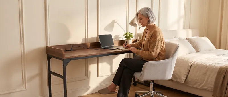 Vrouw zit aan de DreamDesk tegen een lichte wand, met een laptop op het kanteldeel, haar voeten natuurlijk op de vloer en een lamp, notities en water op het blad.