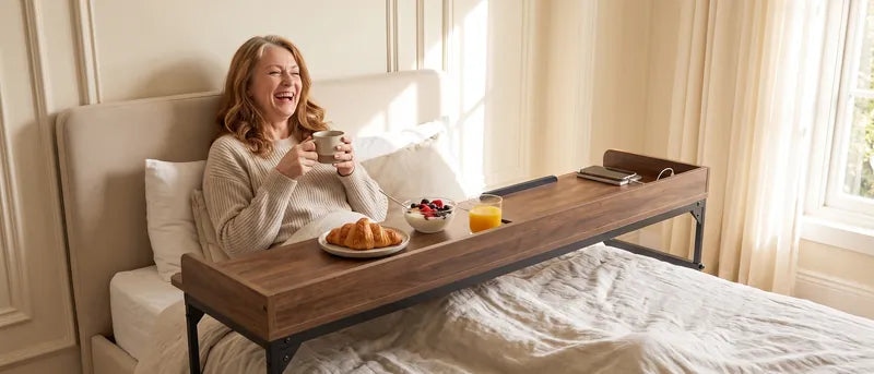 Vrouw ontbijt met een kom en lepel in bed aan de DreamDesk; een koffiemok staat op het blad en haar sokken rusten op het dekbed.