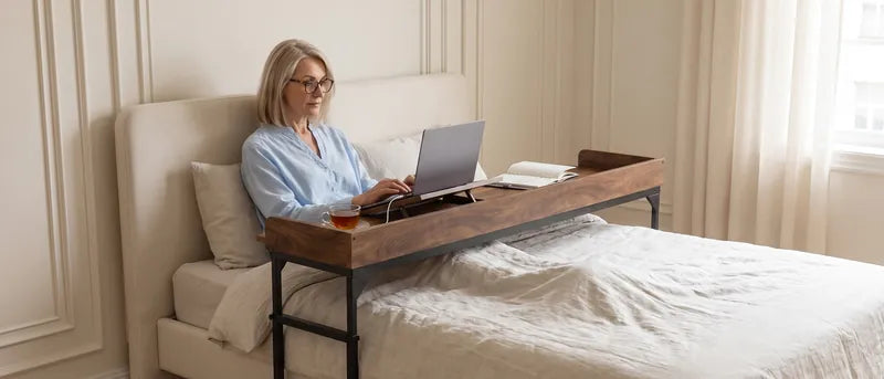 Vrouw zit op bed en gebruikt een laptop op de DreamDesk, met haar benen en sokken op het dekbed.